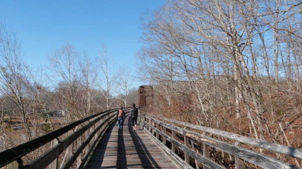 View forward: Two walkers on a boardwalk with converted railroad trestle ahead - among trees and water